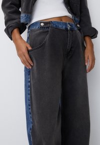 Persona que lleva unos pantalones anchos a dos tonos, con el frente gris oscuro y paneles laterales azules, combinados con una parte superior blanca corta y una chaqueta oscura.