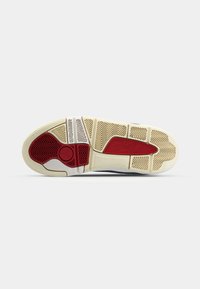 Semelle extérieure de chaussure de sport composée d'un mélange de caoutchouc texturé en rouge, blanc et beige, avec des motifs de crampons distinctifs et un détail de logo circulaire.