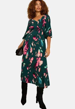 Kleid mit Blumenmuster in Dunkelgrün, mit rosa und türkisfarbenen Akzenten, V-Ausschnitt, lockeren Ärmeln und knielangem Rock. Getragen mit schwarzen Stiefeln.