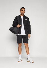 Crna jakna na patentni zatvarač preko bijele majice, uparena s crnim kratkim hlačama. Bijele i crne sportske cipele, nosi crnu torbu za sport.