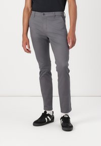 Pantalones gris ajustados de tejido elástico, con frente plano, trabillas para cinturón y corte slim. Combinados con zapatillas negras con acentos blancos.