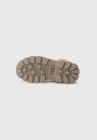 Semelle de chaussure en caoutchouc marron avec des motifs de profondeur, présentant un mélange de motifs circulaires et croisés. Logo de la marque visible dans un accent rectangulaire.