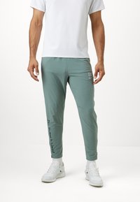 Světlé zelené sportovní kalhoty s tapered střihem, vybavené bočními zipy, bílým prošíváním a nápisem "Žádný člověk není omezen" na levé noze.