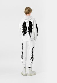 Sudadera blanca con diseño de alas de plumas negras en la parte trasera, combinada con pantalones joggers blancos a juego con detalles laterales. Llevada con botas blancas.