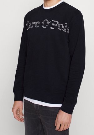 Man draagt een zwarte lange mouw trui met de tekst "Marc O'Polo" op de borst, donkere spijkerbroek en een wit hemd.