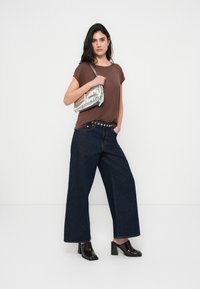 Καφέ κοντομάνικη μπλούζα, ψηλόμεσο παντελόνι από σκούρο τζιν με φαρδιά μπατζάκια, μαύρη ζώνη με καρφιά, ασημένια τσάντα, μαύρα γυαλιστερά παπούτσια με χοντρό τακούνι.