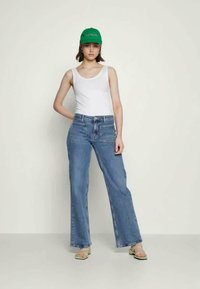 Jeans de perna larga azul claro com dois bolsos frontais, usados com uma camisa de alças branca e um boné verde, emparelhados com sandálias transparentes.