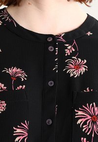 Svart tygtop med rosa blomsterbroderi, rund halsringning och fyra knappar fram. Den har två bröstfickor med blomsterdetaljer.