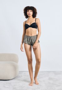 Soutien-gorge noir à armatures et short imprimé léopard avec un design évasé. Le mannequin se tient pieds nus dans un environnement aux tons neutres.