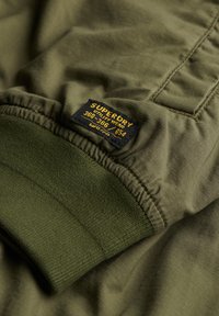 Olive green fabric with textured finish, featuring a ribbed cuff and a rectangular brand label in blue and yellow stitched on it.