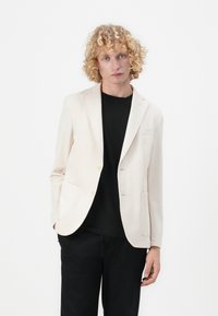 Beige sako s hladkým materiálem, jednobotonové provedení, dvě přední kapsy a slim fit, kombinované s černým tričkem s kulatým výstřihem.