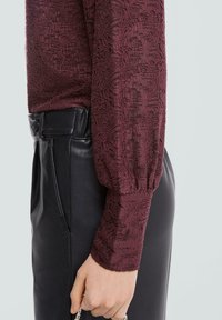Bordowa, fakturowana bluzka z bufionymi rękawami, zestawiona z czarnymi skórzanymi spodniami. Zbliżenie na detale ubrania, ukazujące strukturę tkaniny i dopasowanie.