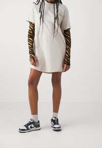 Corto vestido beige con cuello redondo, usado sobre una camiseta de manga larga a rayas en negro y dorado. Combinado con zapatillas negras y blancas con acentos morados.