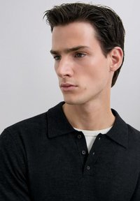 Polo de color gris oscuro hecho de material suave, con un cuello y tres botones en la parte del cuello, superpuesto sobre una camiseta blanca.