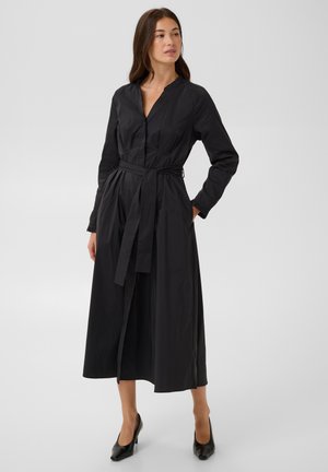 Femme portant une longue robe noire ceinturée à manches longues et des talons noirs, debout avec une main dans la poche, regardant vers la gauche.