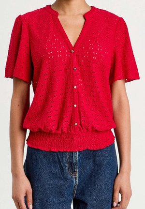 Blouse rouge à manches courtes avec boutons, motif ajouré et taille smockée, portée avec un jean en denim bleu.