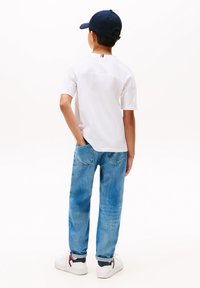 Hvit kortermet skjorte, blå jeans og hvite joggesko med røde og marineblå detaljer; barnet står med hendene i lommene, vendt bort.