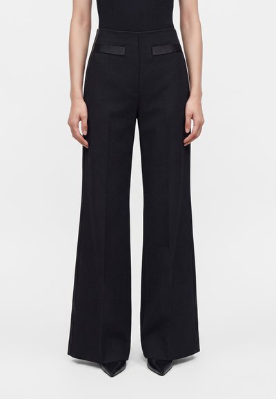 Personne portant un pantalon noir taille haute à jambes larges avec un pli frontal et des poches fendues, associé à des chaussures noires pointues et un haut noir sans manches.