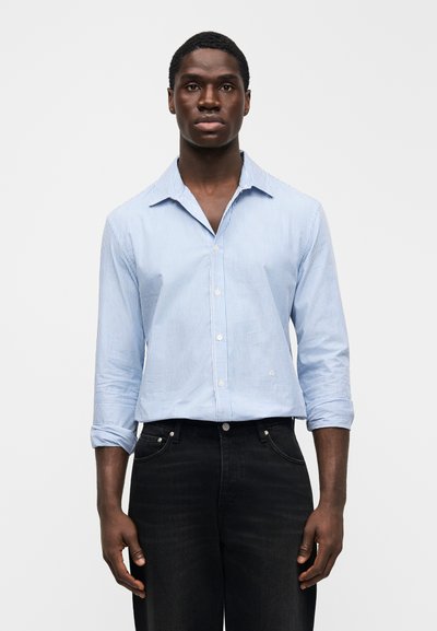 Langærmet skjorte med lyseblå og hvide striber, knaplukning ved kraven, kombineret med sorte jeans; stoffet virker glat og skarpt.