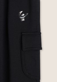 Tissu noir avec un rabat de poche rectangulaire cousu et un logo abstrait argenté brillants au-dessus de la poche sur le vêtement.