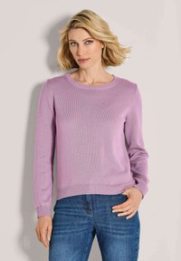 Blonde Frau mit kurzen Haaren, die einen lila Pullover mit langen Ärmeln und Blue Jeans trägt, steht vor einem schlichten hellen Hintergrund.