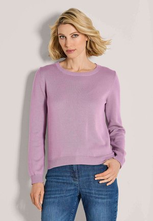 Blonde Frau mit kurzen Haaren, die einen lila Pullover mit langen Ärmeln und Blue Jeans trägt, steht vor einem schlichten hellen Hintergrund.
