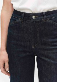 Donkerblauwe spijkerbroek met een taillehoogte midden, voorzien van koperen stiksels, een metalen knoopsluiting en zakken aan de voorkant.