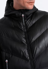 Schwarze Pufferjacke mit Kapuze, ausgestattet mit einem Chevron-Muster, sichtbarem Reißverschluss und Stofftextur. Leichtgewichtig mit glänzendem Finish.