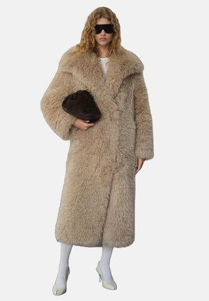 Manteau en fausse fourrure de couleur beige clair, au design surdimensionné avec un large col. Le mannequin tient une pochette marron foncé, portant des collants blancs et des talons clairs.