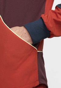 Jachetă roșie și vișinie cu manșete ribbed de nuanță navy, detalii contrastante de crem și o buzunar kangaroo cu o mână parțial vizibilă.