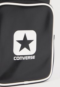 Borsa nera con zip realizzata in materiale liscio, con un grande logo a forma di stella bianca e il testo "CONVERSE", rifinita con dettagli bianchi.