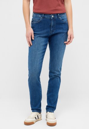 Person trägt blaue Slim-Fit-Jeans, weiße Sneakers mit braunen Sohlen und ein rotes Kurzarmshirt und steht vor einem einfarbigen Hintergrund.
