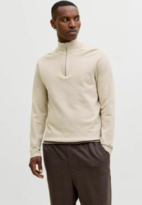 Homme portant un pull à fermeture éclair beige à manches longues et un pantalon à carreaux marron, regardant vers sa gauche avec une main dans sa poche.