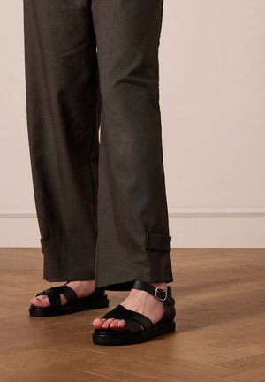 Black sandals with wide straps and a buckle, paired with dark gray wide-leg pants featuring a rolled cuff detail. Smooth texture, casual style.