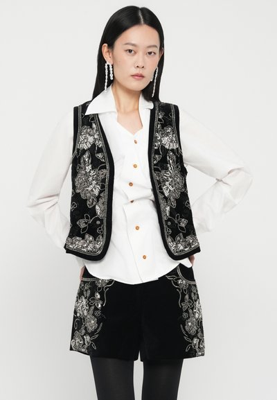 Femeie îmbrăcată într-o bluză albă, vestă din catifea neagră și pantaloni scurți cu broderie florală argintie, purtând cercei lungi din perlă, pozând cu mâinile încadrate pe șolduri.