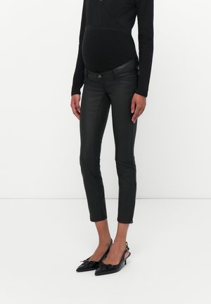Zwangere persoon draagt een zwarte top met lange mouwen, zwarte zwangerschaps skinny broek en zwarte puntige slingback hakken, staand tegen een witte achtergrond.