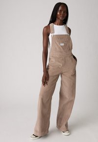 Tan corduroy overalls with wide legs, adjustable straps, and front pocket detail. Worn over a white fitted top, paired with black sneakers.