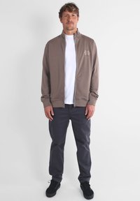 Sweatshirt zippé marron avec col montant et broderies florales, porté sur un t-shirt blanc. Assorti avec un pantalon gris foncé et des baskets noires.