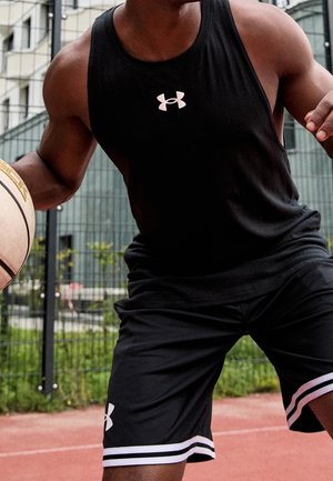 Athlet in schwarzem Under Armour Tanktop und Shorts, der auf einem Outdoor-Platz mit Metallzaun und urbanem Hintergrund einen Basketball dribbelt.