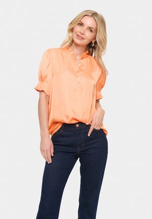 Persikofärgad satinblus med vågig krage, korta puffärmar och knappslå. Matchad med mörkblå jeans med orange sömmar.