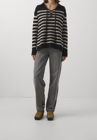 Schwarz-beige gestreifter Pullover mit Kragen und Schnürdetails, kombiniert mit grauen Jeans und olivgrünen Plateauschuhen.