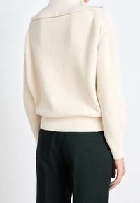 Osoba ubrana w beżowy sweter z dzianiny o szerokim kołnierzu i czarne spodnie, pokazana od tyłu na jednolitym tle.