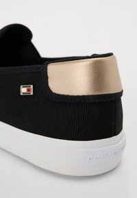 Svart tygsneaker med en guldfärgad metallic hälackent. Har en vit gummisula och en liten logotags i blått, rött och vitt.