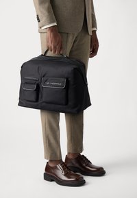 Sac de voyage en nylon noir avec deux poches frontales, logo, poignée supérieure et fermeture éclair. Porté avec un costume beige et des chaussures marron.