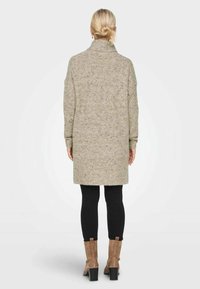 Langer, hellgrauer Strickpullover mit hohem Kragen und gerippter Textur, getragen über schwarzen Leggings und beigen Knöchelstiefeln.