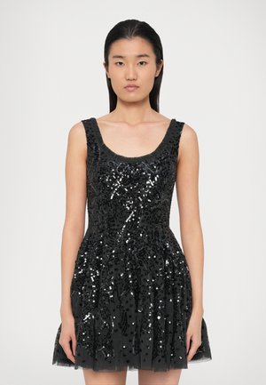 Femme aux cheveux noirs raides portant une robe noire sans manches ornée de sequins noirs dans un motif floral sur un fond clair uni.