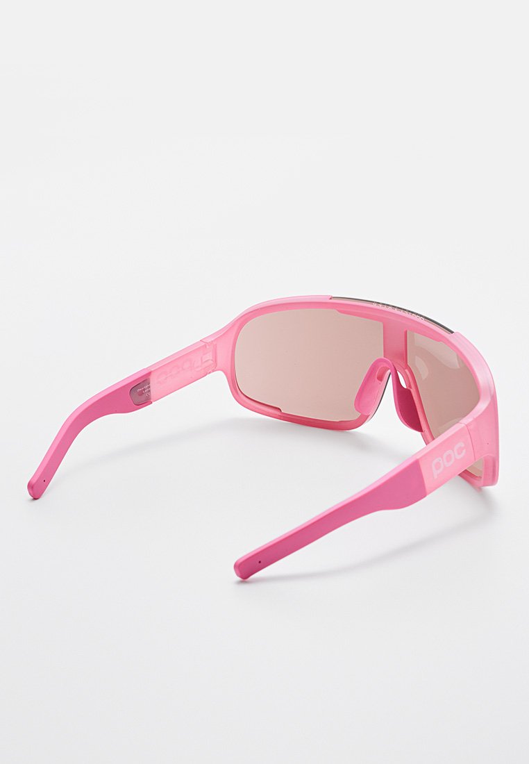 Jasnoróżowe okulary przeciwsłoneczne z okrągłym kształtem, przyciemnionymi soczewkami i teksturowanym wykończeniem. Posiadają logo "POC" na boku oprawki.