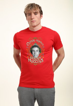 Mladý muž s blond vlasy nosící červené tričko s černobílou tváří a nápisem "Jsem bavlněný hloupý brouk" v barevném designu.