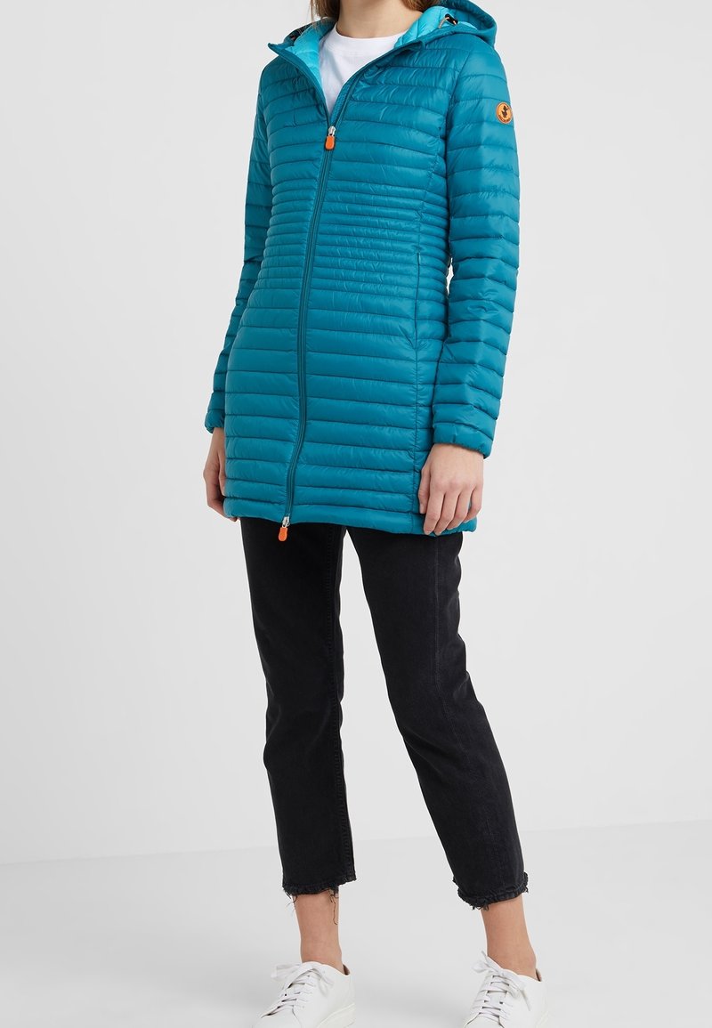 Abrigo acolchado en color azul verdoso con capucha y tiradores de cremallera naranjas, combinado con jeans negros cortos y zapatillas blancas, frente a un fondo liso.