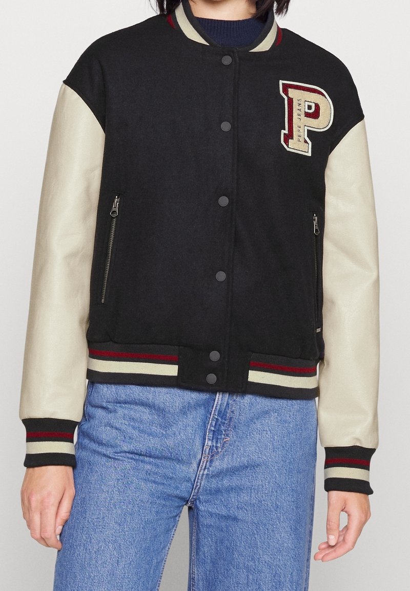 Personne portant une veste universitaire noire et beige avec des poches zippées et un grand écusson en forme de "P" sur la poitrine, associée à un jean bleu.